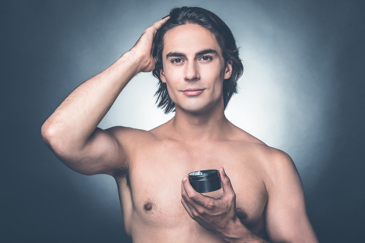 Portrait of young shirtless man looking at camera while applying hair gel and standing against grey background